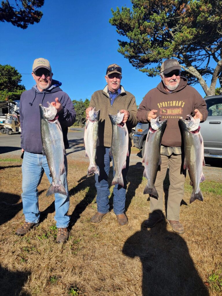 Three anglers display six fresh-caught chinook salmon in parking area with trees and blue sky behind them.