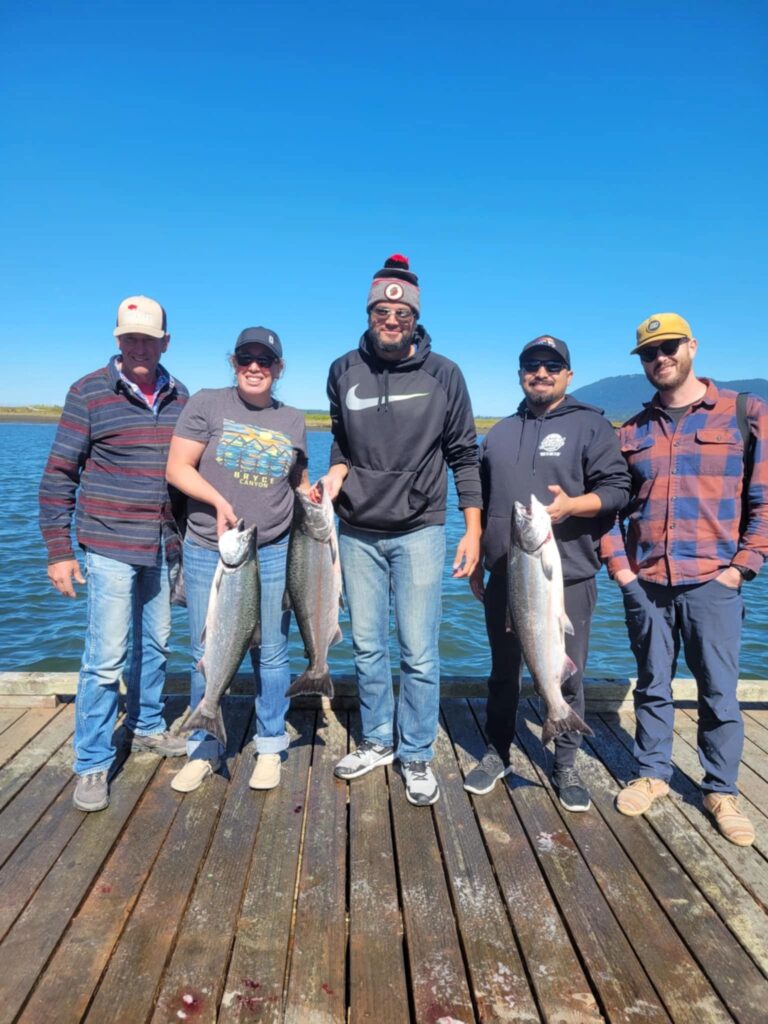 Five anglers on wooden dock displaying five chinook salmon with calm bay waters and mountains behind