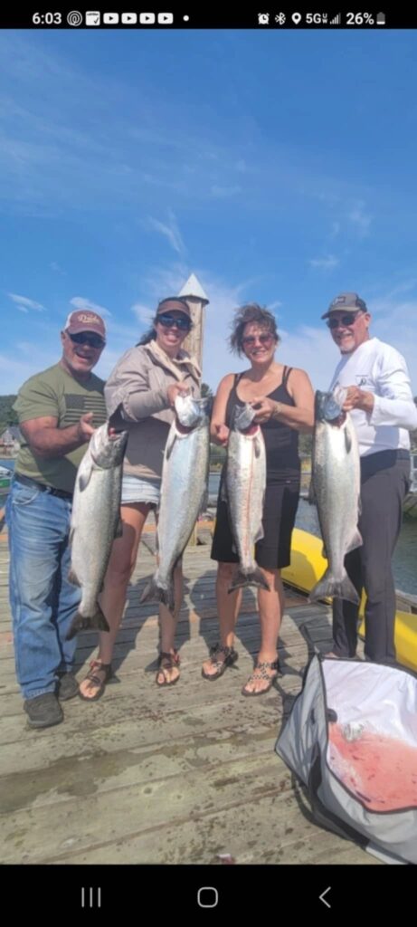 Four anglers on dock holding chinook salmon with lighthouse visible in background under blue sky