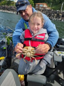 Adult and child in life jacket holding Dungeness crab on boat with crab pots and rocky shore visible