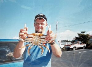 Man in blue tank top holding large Dungeness crab in parking lot with vintage trucks in background
