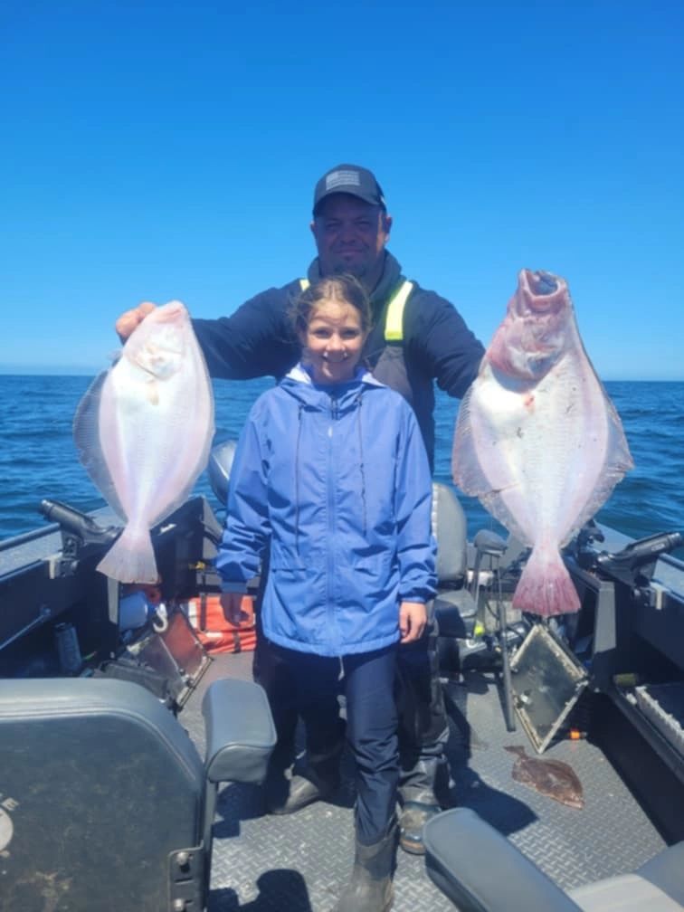 Man and child on boat holding two Pacific halibut under clear blue sky on ocean
