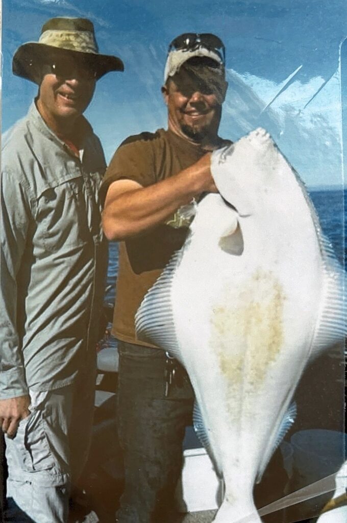 Two anglers on boat holding large halibut with white underside visible against blue ocean water