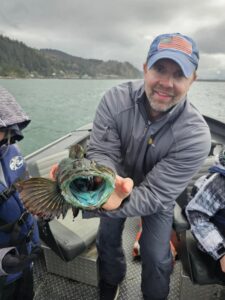 Angler in boat holding lingcod with bright blue-green mouth open, forested hills and water in background