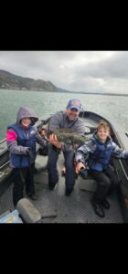 Adult and two children in life jackets holding lingcod on aluminum boat in coastal waters