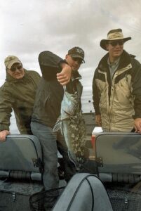 Three anglers on boat displaying large lingcod catch under overcast sky on ocean water