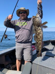 Angler on boat holding large lingcod by jaw with fishing rod in other hand on calm ocean water.
