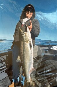 Angler on boat holding large lingcod by mouth with calm bay and hills in background under blue sky
