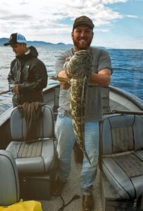 Angler holding large lingcod on boat with mountains and water visible in background