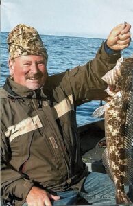 Angler in camouflage cap holding large spotted lingcod on boat with calm ocean water behind