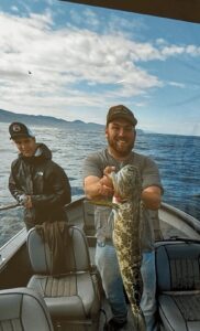Angler holding large lingcod on boat with mountains visible across water under cloudy sky