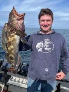 Angler on boat holding large lingcod with mouth open in ocean waters under blue sky