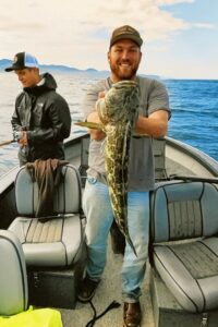 Angler holding large lingcod on boat with mountains and calm water in background