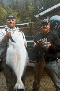 Two anglers displaying halibut and lingcod catches in front of boat and truck in forested area