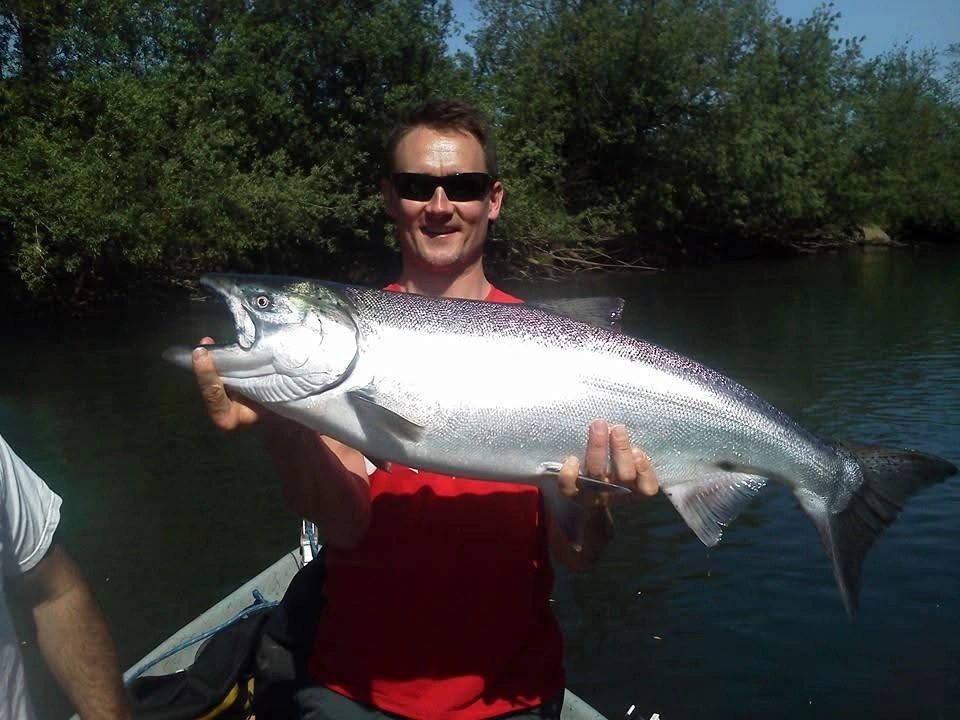 Angler in sunglasses holding large chinook salmon on boat in calm river with forested shoreline.