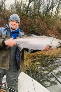 Angler in gray beanie and waders holding large chinook salmon at river's edge with bare branches behind