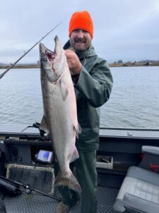 Angler in bright orange beanie holding large chinook salmon on boat with flat wetland landscape behind