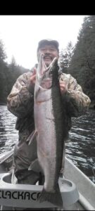 Angler in camouflage jacket holding large chinook salmon while standing in boat on forested river