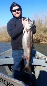 Angler in black hoodie and sunglasses holding large chinook salmon on small fishing boat in river.