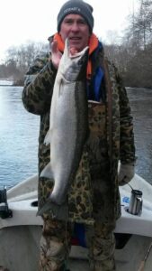 Angler in camouflage holding large chinook salmon on boat in winter river setting with bare trees.