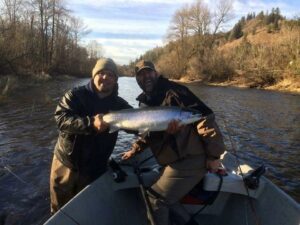 Two anglers in drift boat holding large chinook salmon on winter river with forested hills