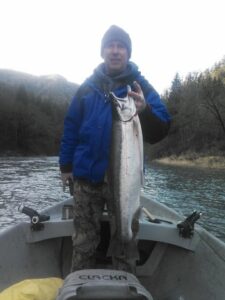 Angler in blue jacket holding large chinook salmon in aluminum drift boat on forested river
