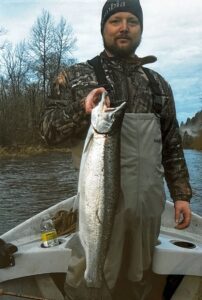Angler in camouflage waders holding large chinook salmon in aluminum boat on river with bare trees