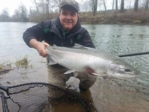Angler in waders holding large chinook salmon while kneeling in shallow water on misty winter day