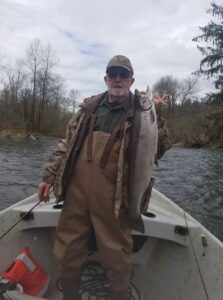 Angler in camouflage jacket holding large chinook salmon on small boat in river with bare trees.