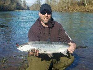 Angler in brown hoodie displaying large chinook salmon at winter riverbank with flowing water behind.