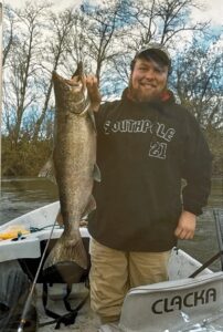 Angler in boat holding large chinook salmon on winter river with bare trees in background.