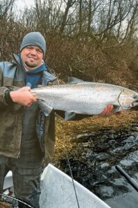 Angler in waders holding large chinook salmon at riverbank with bare winter vegetation behind