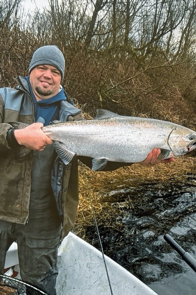 Angler in waders holding large chinook salmon at riverbank with bare winter vegetation behind