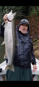 Angler in navy rain jacket holding large chinook salmon vertically on boat in forested river setting