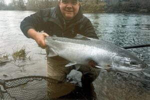 Angler kneeling in shallow water holding large chinook salmon with net beside him on riverbank