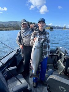 Young angler holding large chinook salmon on boat with adult companion, calm river and forested hills behind