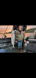 Child holding large chinook salmon on aluminum fishing boat at sunset with forested hills in background