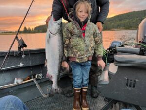 Young child holding large chinook salmon on boat at sunset with forested hills in background