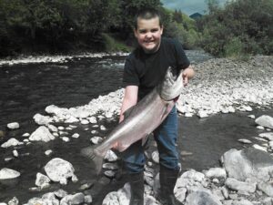 Young angler displays large chinook salmon while standing in shallow rocky river in Oregon.