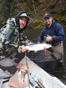 Two anglers in boat holding coho salmon with fishing net and gear visible on wooded river