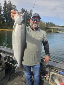 Angler in sunglasses holding large coho salmon on aluminum fishing boat with bridge and evergreen trees visible
