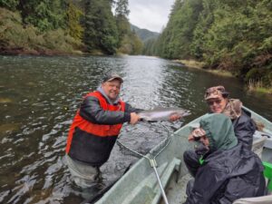 Two anglers in drift boat display silver salmon with fishing net on forested river