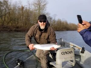 Angler in brown jacket holding silver coho salmon in drift boat on river with bare trees in background