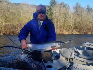 Angler in blue jacket holding silver coho salmon while kneeling in drift boat on river.