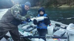Adult and child in winter clothing hold coho salmon on drift boat on misty river
