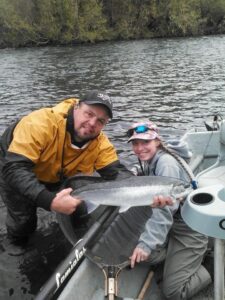 Man and child in boat holding coho salmon on river with forested shoreline