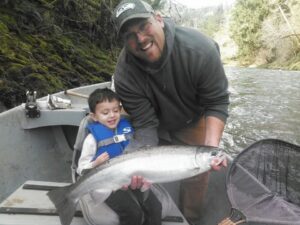 Man and young child in blue life vest holding coho salmon in drift boat on river