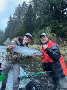 Two anglers in boat displaying a bright silver coho salmon with forested riverbank behind them.