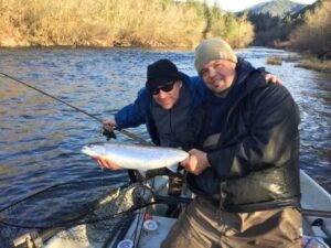Two anglers in a drift boat displaying a silver salmon caught on a river in winter.