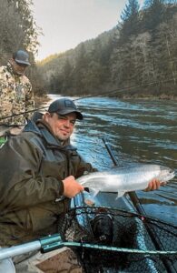 Angler in drift boat holding fresh coho salmon on forested river with guide in background.